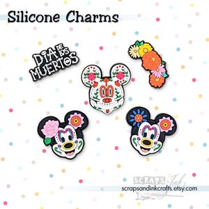 Peut inclure: Charm en silicone avec des motifs du Jour des Morts. Comprend un charme texte "Dia de los Muertos" et des breloques tête de Mickey Mouse décorées de fleurs et de motifs de têtes de mort. Les breloques sont colorées et détaillées.