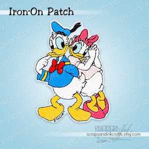 Può includere: Toppa termoadesiva ricamata con Paperino e Paperina. Paperino indossa una camicia e un cappello da marinaio blu, con un papillon rosso. Paperina indossa un fiocco e un vestito rosa. La toppa ha il testo "Iron-On Patch".