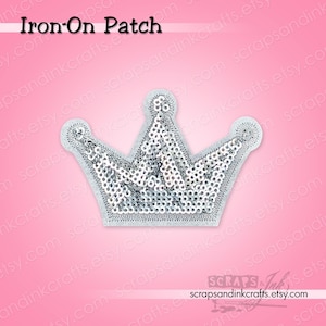 Könnte beinhalten: Silberner Pailletten-Kronen-Aufnäher mit weißem Rand. Der Aufnäher hat ein detailliertes Design und befindet sich auf einem rosa Hintergrund. Der Text "Iron-On Patch" steht oben.