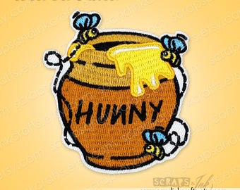 May include: Embroidered iron-on patch depicting a brown honey pot with the word "HUNNY" and yellow honey dripping from the top. Three bees with blue and yellow details surround the pot. The patch has a white border and the text "Iron-On Patch".