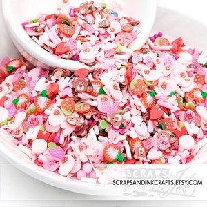 May include: A bowl filled with pink, white, and red sprinkles. The sprinkles are shaped like strawberries, hearts, bears, and stars. The sprinkles are perfect for decorating cakes, cupcakes, and other desserts.