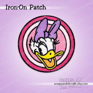 Può includere: Toppa rotonda termoadesiva con Daisy Duck. Il design presenta uno sfondo a righe rosa e bianco, un bordo rosa e il volto di Daisy Duck. Il testo "Iron-On Patch" è in alto. Adatta per abbigliamento e accessori.