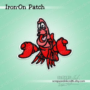 May include: Embroidered iron-on patch featuring a cartoon crab in red and pink with a white border. The patch has the words "Iron-On Patch" at the top. The crab has a smiling expression and is a fun accessory for clothing or bags.