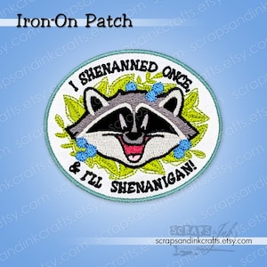 May include: A circular embroidered iron-on patch featuring a raccoon face surrounded by green leaves and small blue flowers. The text reads I SHENANNED ONCE, & I'LL SHENANIGAN! The patch has a light blue border and a white background with black stitching.