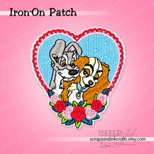 May include: Embroidered iron-on patch featuring two dogs in a heart-shaped design. The patch has a light blue background with a red scalloped border. The dogs are surrounded by a floral arrangement of red and pink flowers. The text "Iron-On Patch" is at the top.