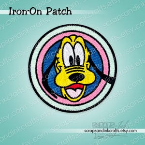 Può includere: Toppa termoadesiva ricamata con Pluto, il cane dei cartoni animati Disney, in un design circolare. La toppa ha un viso giallo, orecchie nere e una lingua rossa, su sfondo blu e rosa. Il testo "Iron-On Patch" è in alto.