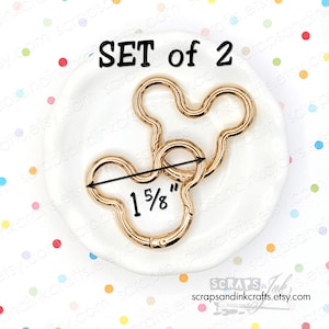 Könnte beinhalten: Set aus zwei goldfarbenen Mickey-Mouse-förmigen Drehverschlüssen, jeweils ca. 4,1 cm groß. Die Verschlüsse liegen auf einer weißen Schale. Der Text "SET of 2" ist sichtbar.