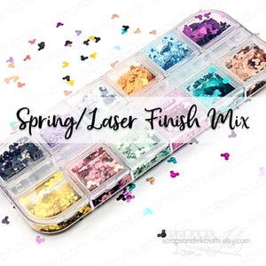 May include: A clear plastic organizer filled with various colors of glitter in a Mickey Mouse shape. The text "Spring/Laser Finish Mix" is displayed. Colors include gold, pink, blue, and purple. Scattered glitter pieces are also visible.