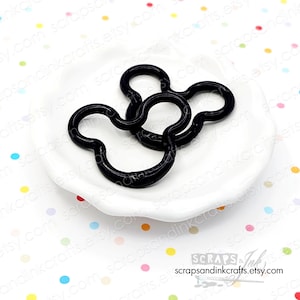 May include: Two black Mickey Mouse-shaped charms rest on a white, scalloped dish. The charms are glossy and have a smooth finish. The background is white with colorful polka dots. The Etsy shop name is visible in the bottom right corner.