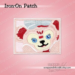 May include: Embroidered iron-on patch featuring a cartoon bear face with a light pink and cream color scheme. The bear has blue eyes, a black nose and mouth, and a light blue bow. The patch is rectangular with the text "Iron-On Patch".