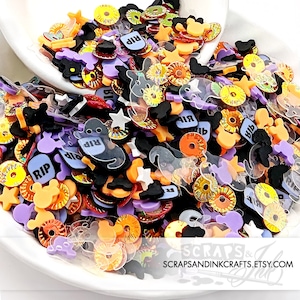 May include: A bowl filled with colorful Halloween-themed resin embellishments. The embellishments include ghosts, bats, stars, Mickey Mouse heads, and rings. Some of the embellishments have the word "RIP" written on them.