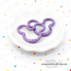 Könnte beinhalten: Zwei lavendelfarbene, Mickey Mouse-förmige Drehverschlüsse. Die Verschlüsse sind aus Metall und haben einen federbelasteten Mechanismus. Sie liegen auf einer weißen Schale mit gewelltem Rand. Der Etsy-Shop-Name "scrapsandinkcrafts.etsy.com" ist sichtbar.