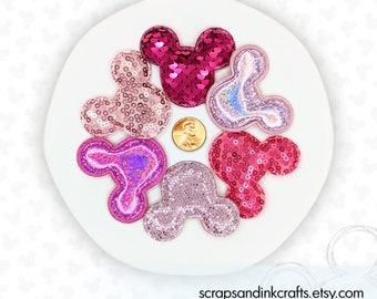 Könnte beinhalten: Sechs rosa, violette und schimmernde Glitzer-Mickey-Maus-Kopfformen. Die Formen bestehen aus Pailletten und sind auf einem weißen Teller in einem Kreis angeordnet.