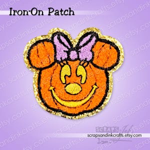 May include: An iron-on patch featuring a pumpkin shaped like a mouse head with large round ears, a yellow nose, and a wide yellow smile. The patch has a light purple bow between the ears and is outlined with a shimmering gold glitter border.