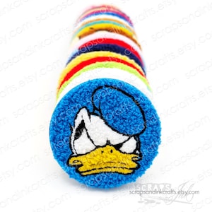 May include: A stack of colorful fabric circles with embroidered cartoon characters. The top circle is blue with a white and yellow embroidered cartoon duck with an angry expression.