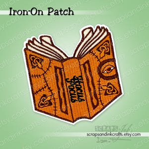 May include: Embroidered iron-on patch featuring an open book with the words "Hocus Pocus" in black. The book is orange with brown stitching and details, including an eye and other symbols. The patch has a white border.