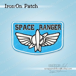 May include: A blue and white embroidered Space Ranger iron-on patch. The patch features the words "SPACE RANGER" in black letters above a white and black emblem of wings and a rocket. The patch is rectangular with rounded corners.