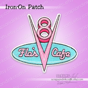 Può includere: Una toppa ricamata da stirare con un design a motore V8 rosa e bianco e la scritta "Fla's Cafe" in un font retrò. La toppa ha uno sfondo ovale azzurro chiaro e un bordo bianco. Le parole "Iron-On Patch" sono in alto.