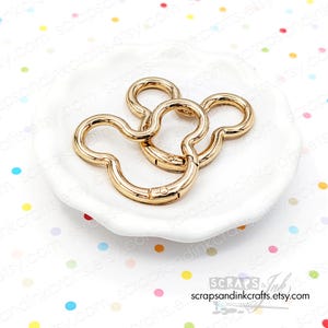 May include: Gold-colored metal Mickey Mouse-shaped swivel clasps. The clasps are on a white, scalloped-edge dish. The background is white with colorful polka dots. The Etsy shop name, scrapsandinkcrafts.etsy.com, is visible in the bottom right corner.