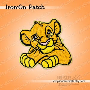 May include: An embroidered iron-on patch featuring a yellow lion cub with large eyes, black outlines, and a pink nose. The patch is set against an orange background with the text Iron-On Patch at the top and scrapsandinkcrafts.etsy.com at the bottom.