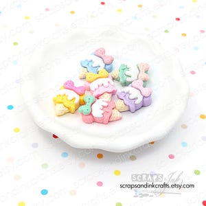 May include: A collection of colorful ice cream cone charms in various pastel colors, including yellow, pink, blue, green, and purple, with white frosting and bow accents. The charms are arranged on a white, slightly scalloped dish.