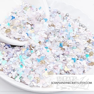 May include: A close-up of a mix of craft embellishments in a white bowl. The mix includes white, iridescent, and light blue Mickey Mouse-shaped sequins, small pearls, and gold-colored beads. The Etsy shop name, SCRAPSANDINKCRAFTS.ETSY.COM, is visible.
