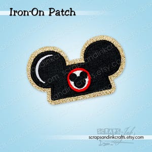May include: Black iron-on patch in the shape of a classic movie camera with a gold glitter border. The patch features a red circle with a white Mickey Mouse head design. The text "Iron-On Patch" is at the top.