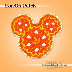 May include: An orange iron-on patch in the shape of a mouse head. The patch has a gold glitter border, yellow and white accents, and the text "Iron-On Patch" at the top. The patch is likely for clothing or accessories.