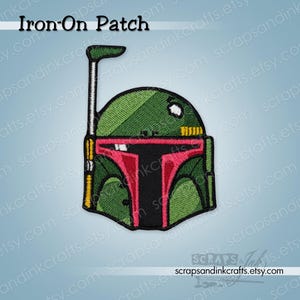May include: An embroidered iron-on patch featuring a stylized green helmet with a black T-shaped visor, a red border around the visor, and a thin antenna extending from the left side. The text Iron-On Patch is visible at the top of the image.