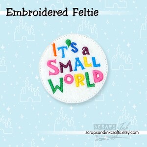 Może przedstawiać: Haftowany filc z napisem "It's a Small World" w kolorowych literach. Filc jest biały i okrągły, z obszytą krawędzią. Tło jest jasnoniebieskie z wzorami zamków i gwiazd.