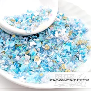May include: A white bowl filled with blue and white sprinkles, beads, and other decorative elements. The sprinkles are in a variety of shapes, including stars, hearts, and circles. There are also some gold sprinkles mixed in. The bowl is sitting on a white surface. The text "Scraps & Ink" is visible on the bottom right corner of the image.