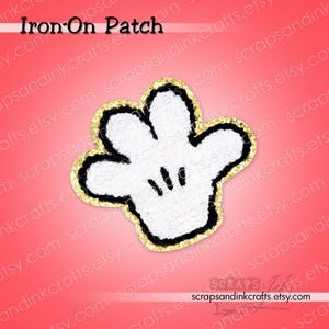 May include: A cartoon hand-shaped iron-on patch, featuring a white palm with black detailing and a gold glitter border. The text "Iron-On Patch" is at the top. The patch is designed for fabric application.