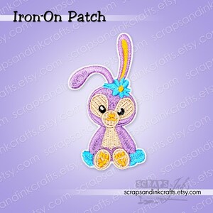 May include: An embroidered iron-on patch featuring a purple rabbit with long ears, a light tan face and belly, and yellow paw pads. The rabbit has a bright blue flower on its head and blue accents on its feet, all outlined in white thread.