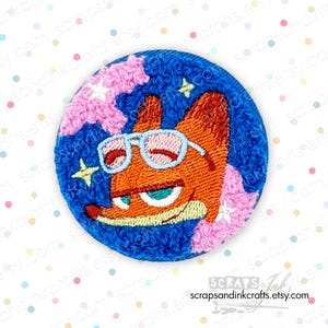 May include: Embroidered patch showing an orange fox wearing sunglasses, set against a blue background with pink accents and stars. The patch has a textured appearance and the text "SCRAPS & Ink" is visible.