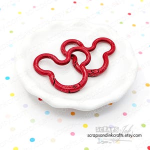 May include: Two red Mickey Mouse-shaped swivel clasps are displayed on a white, scalloped dish. The clasps are made of metal and have a glossy finish. The background is white with colorful polka dots. The Etsy shop name is visible.