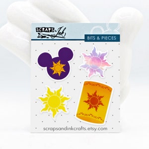 Puede incluir: Cuatro pegatinas que representan una cabeza de Mickey Mouse morada con un sol amarillo en el interior, un sol amarillo con rayos naranjas, un sol morado y rosa con rayos naranjas y una linterna amarilla con un sol rojo en el interior. Las pegatinas están en una hoja blanca con el texto "Scraps & Ink" y "Bits & Pieces" impreso en ella.