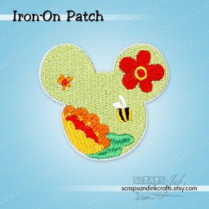 May include: An iron-on patch in the shape of a mouse head, featuring a light green background with embroidered details. The design includes a red flower, a bee, and other floral elements. The text "Iron-On Patch" is at the top.