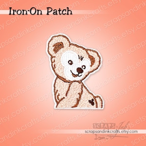 May include: An embroidered iron-on patch featuring a light brown teddy bear with a white face, dark brown nose, and a small dark heart shape on its hip. The text Iron-On Patch is visible at the top, and the brand name is at the bottom.