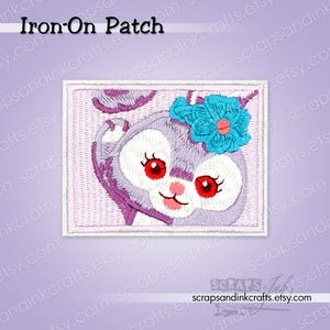 May include: An embroidered iron-on patch featuring a cartoon bunny with large red eyes, a pink nose, and a blue flower. The patch has a light purple background and the text "Iron-On Patch" at the top.
