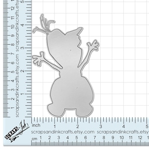 May include: A silver metal die cut of a snowman with a carrot nose and stick arms. The die cut is approximately 2 inches tall and is shown on a grid background with a ruler for scale.
