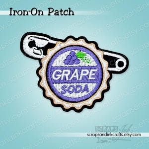 May include: Embroidered iron-on patch featuring a purple and white "Grape Soda" design with a cluster of grapes. The patch is shaped like a bottle cap and is attached to a black and white safety pin. The text "Iron-On Patch" is at the top.