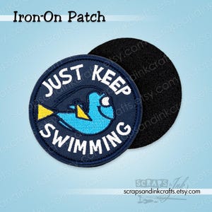 May include: Round iron-on patch with a blue and yellow cartoon fish design. The patch has the words "JUST KEEP SWIMMING" in white lettering on a navy blue background. A black felt backing is also visible.