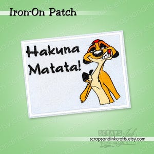 May include: Rectangular iron-on patch with the words "Hakuna Matata!" in black on a white background. It features a cartoon meerkat in orange, tan, and black. The patch has a white border and is designed for fabric application.