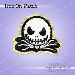 May include: An iron-on patch featuring a stylized skull with a wide grin and black eyes, outlined in black and white. The patch has a gold glitter border and the text "Iron-On Patch" at the top.