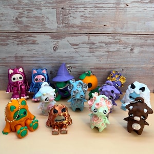 May include: A collection of colorful, small figurines with various designs, including a cat, a witch, a pumpkin, and other fantasy creatures. Each figure has a keychain attached, and they are displayed on a light-colored surface.