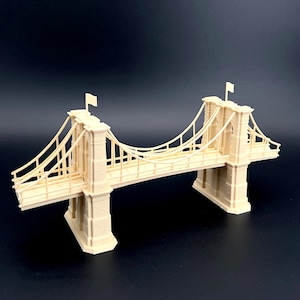 May include: A beige model of a suspension bridge with two towers, a roadway, and suspension cables. The bridge has two small flags on top of the towers. The model is made of a light-colored material and is set against a dark background.