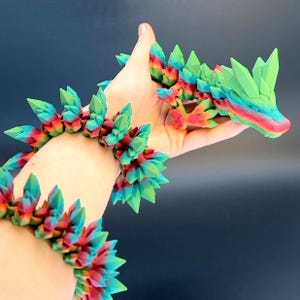 May include: A colorful, 3D-printed dragon sculpture and matching bracelet. The dragon is held in a hand, showcasing its vibrant green, red, and blue hues. The bracelet, with similar crystal-like spikes, wraps around the wrist, creating a cohesive set.