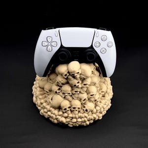 May include: A white and black video game controller rests on a beige skull-shaped base. The controller has black buttons and directional pads. The base is a detailed sculpture of numerous skulls, creating a unique gaming accessory.