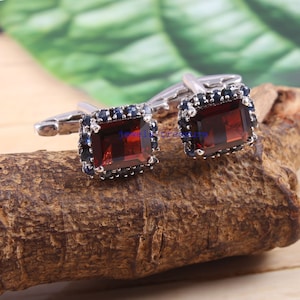 May include: A pair of silver cufflinks featuring a square red gemstone set within a halo of smaller blue gemstones.