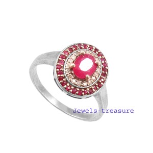 Op de afbeelding: Een zilveren ring met een grote ovale robijn in het midden, omringd door twee concentrische cirkels van kleinere rode en heldere stenen. De ring heeft een eenvoudige band. De tekst "Jewels-treasure" is zichtbaar.